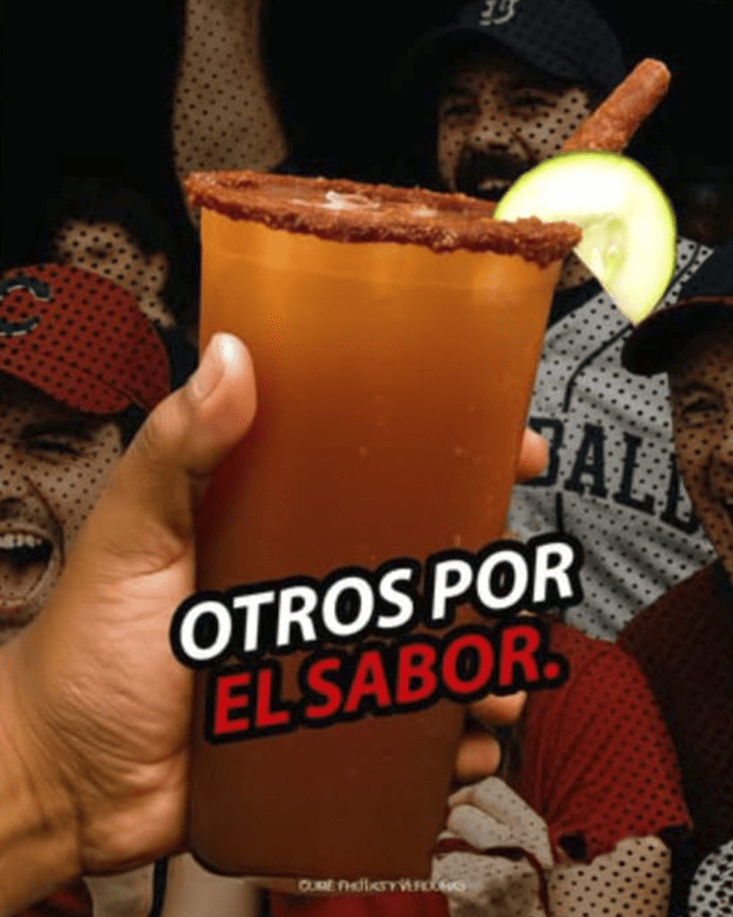 Vaso de michelada o clamato preparado con chile en polvo y una rodaja de pepino, sostenido por una mano en un estadio de béisbol o evento deportivo. El texto en la imagen dice "OTROS POR EL SABOR".