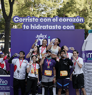 Fotografía de una corredora participando en la 13ª Carrera Kardias TUDN, portando una playera con el logo de Jumex Mia Agua Purificada. En primer plano se observa un banderín morado promocional con el mensaje "5 KM CORRAMOS JUNTOS EN 30 MIN. ¡SÍGUEME!" y logotipos de los aliados de la campaña "Hidratación con Causa".