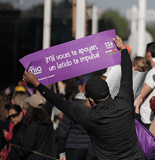 Un espectador sostiene un letrero morado durante la 13ª Carrera Kardias TUDN, con el mensaje motivador: "¡Mil voces te apoyan, un latido te impulsa!". El letrero incluye los logotipos de Jumex Mia y la fundación Kardias, destacando el apoyo de la marca en eventos deportivos con causa social.