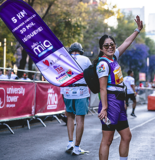 Fotografía de una corredora participando en la 13ª Carrera Kardias TUDN, portando una playera con el logo de Jumex Mia Agua Purificada. En primer plano se observa un banderín morado promocional con el mensaje "5 KM CORRAMOS JUNTOS EN 30 MIN. ¡SÍGUEME!" y logotipos de los aliados de la campaña "Hidratación con Causa".