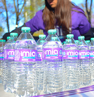 Múltiples botellas de Jumex Mia Agua Purificada de 500 ml dispuestas en una mesa para un evento de hidratación. Las etiquetas moradas con el eslogan "CON VOZ PROPIA" son claramente visibles, mientras al fondo se aprecia a una persona con el uniforme de la marca organizando la distribución.